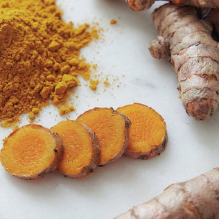 Sliced turmeric rounds with loose turmeric powder and fresh ginger on a white surface.