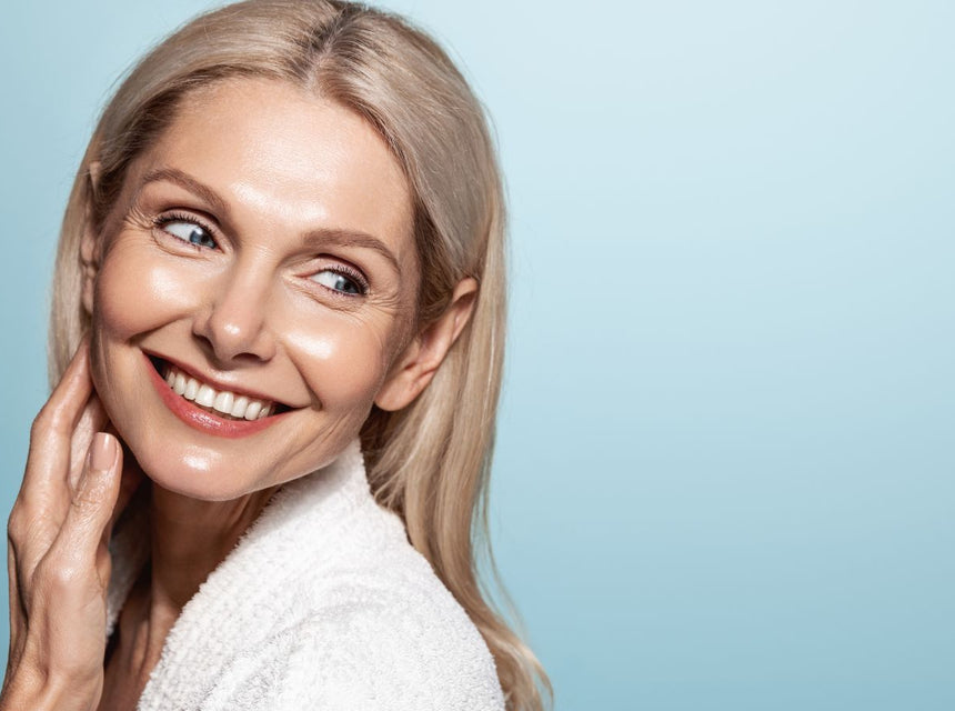 Smiling mature woman with blonde hair touching her cheek against a soft blue backdrop