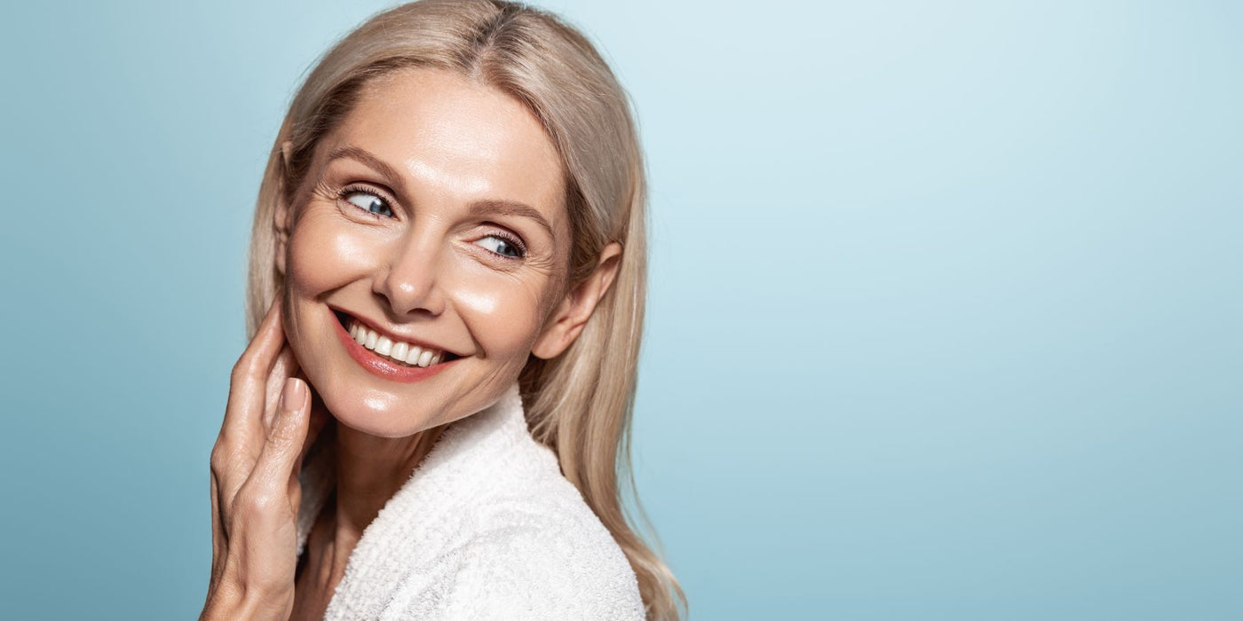 Smiling mature woman with blonde hair touching her cheek against a soft blue backdrop