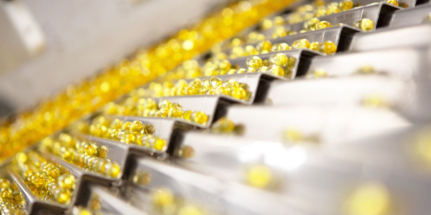 Rows of gold softgel capsules on a conveyor in a supplement production line.