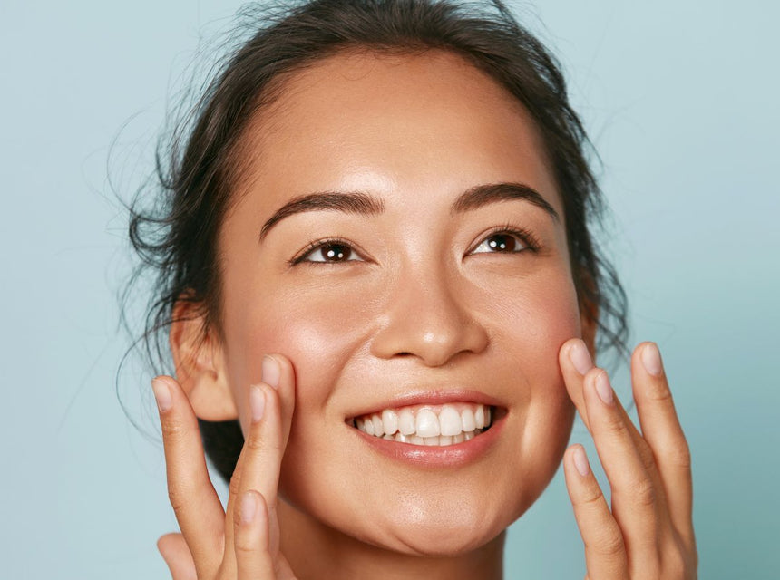 Smiling woman with clear glowing skin touching her cheeks against a soft blue background