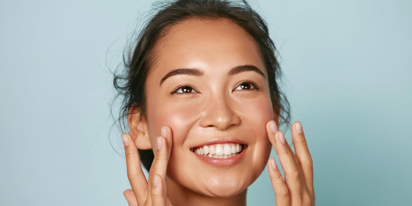Smiling woman with clear glowing skin touching her cheeks against a soft blue background