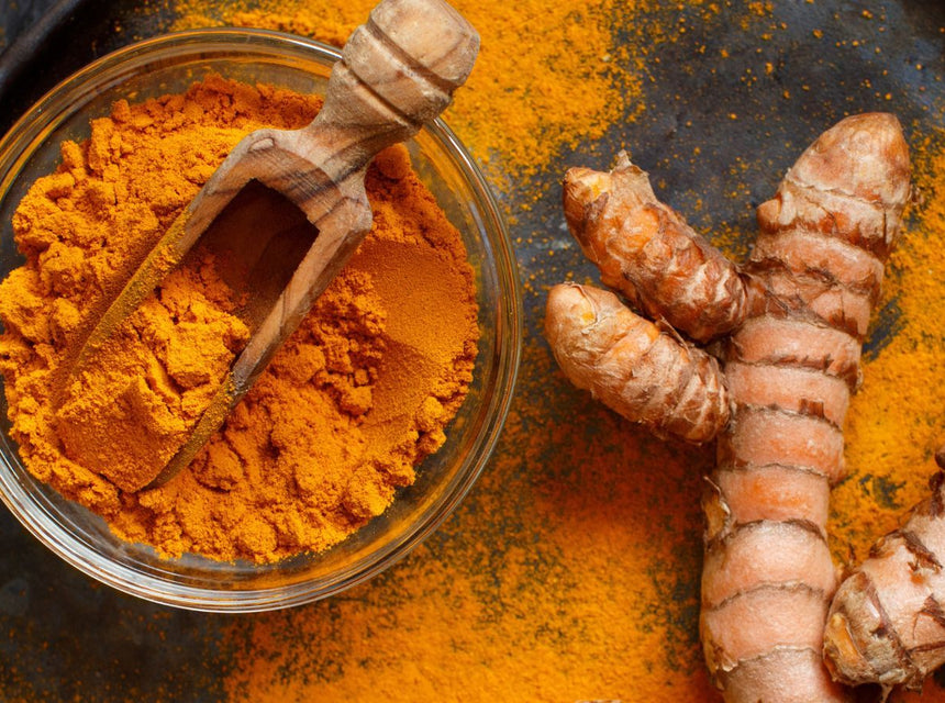 Glass bowl of turmeric powder with a wooden scoop and fresh turmeric roots on a dark textured surface.