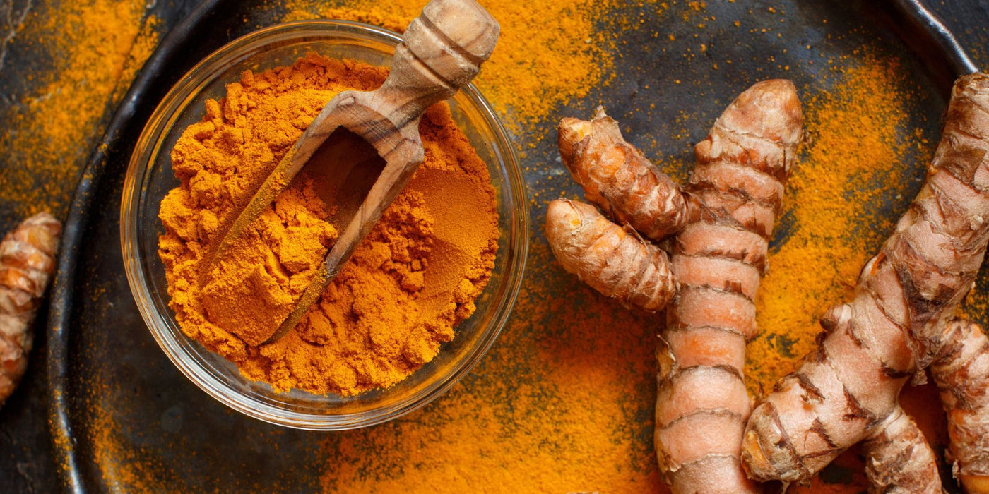 Glass bowl of turmeric powder with a wooden scoop and fresh turmeric roots on a dark textured surface.