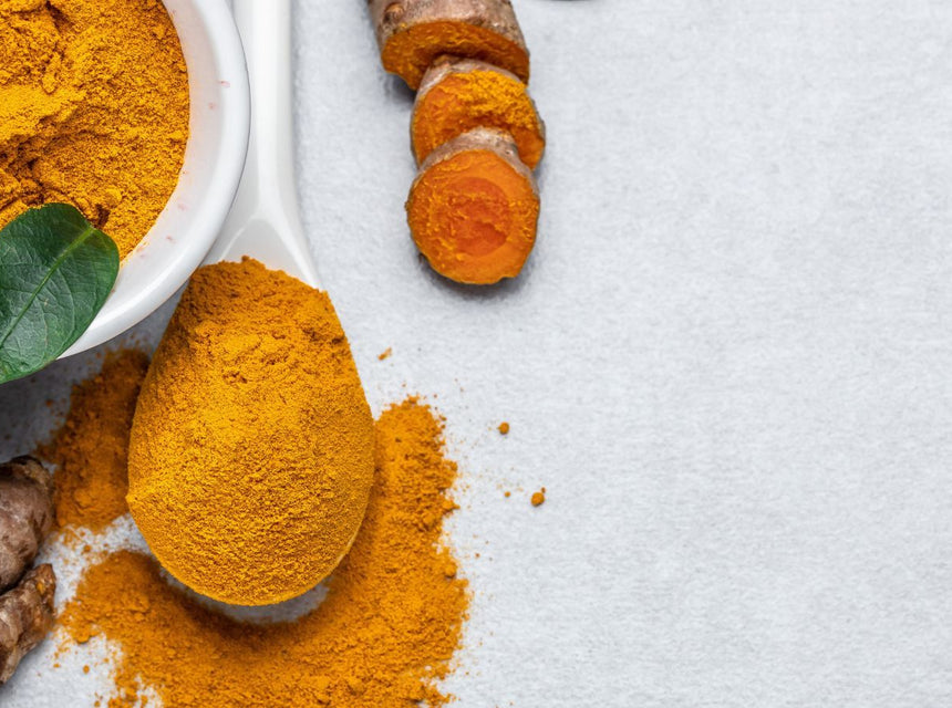 Bright turmeric powder in a white bowl with a fresh green leaf and turmeric roots on a light surface