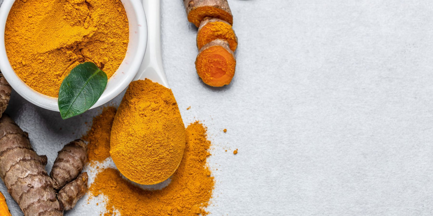Bright turmeric powder in a white bowl with a fresh green leaf and turmeric roots on a light surface