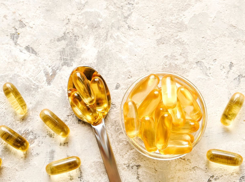 Golden omega-3 capsules in a glass bowl with a spoon on a light textured surface