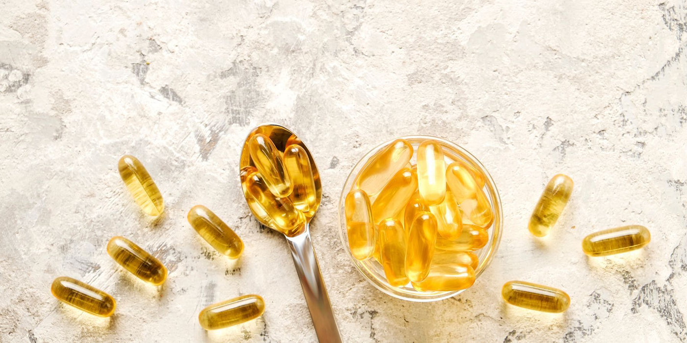 Golden omega-3 capsules in a glass bowl with a spoon on a light textured surface