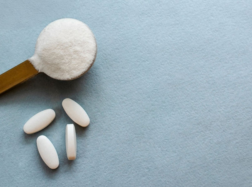 Wooden scoop heaped with white collagen powder and a few white tablets on a pale blue surface