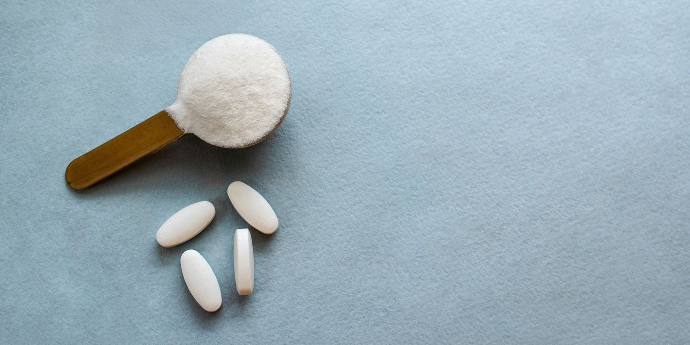Wooden scoop heaped with white collagen powder and a few white tablets on a pale blue surface