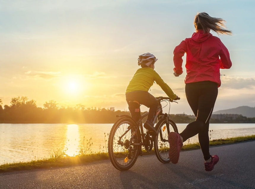 Parent jogging beside child riding a bicycle along a lakeside path at sunset