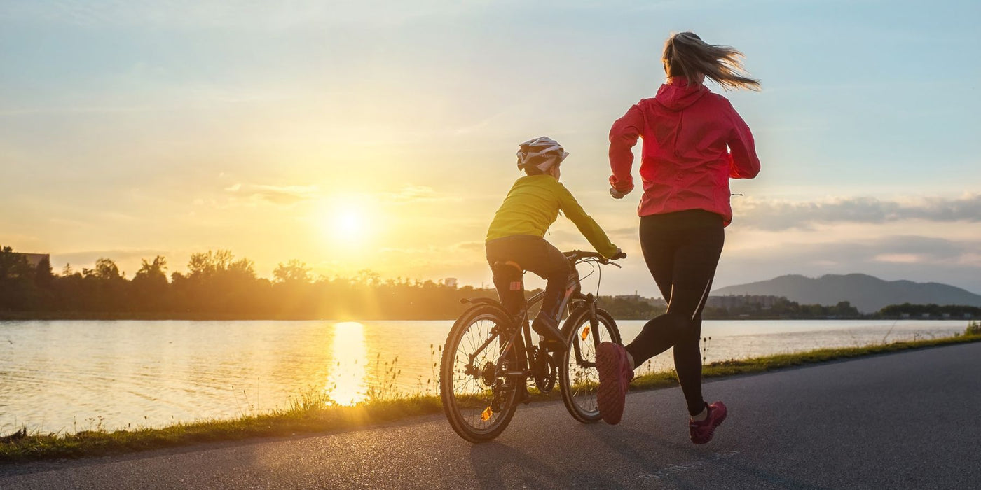 Parent jogging beside child riding a bicycle along a lakeside path at sunset