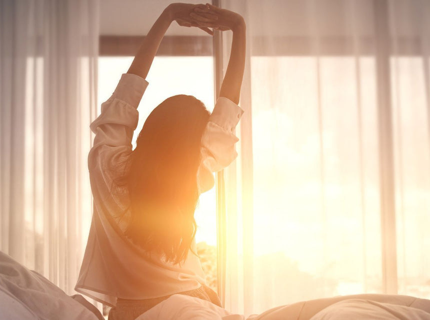 Person stretching arms overhead by a sunlit window in a cozy bedroom.