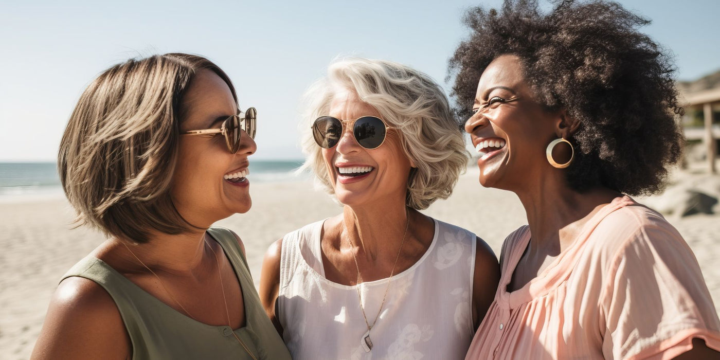 Three women wearing sunglasses laugh together on a sunny beach