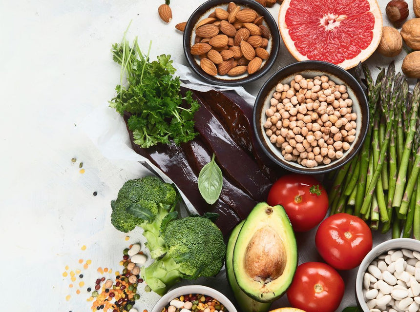 Assorted folate rich foods including avocado, tomatoes, greens and nuts on a light background.
