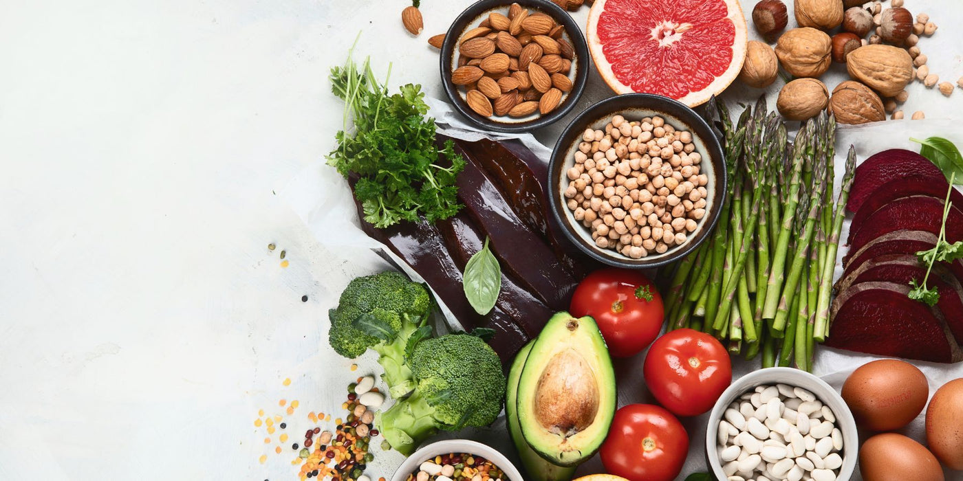 Assorted folate rich foods including avocado, tomatoes, greens and nuts on a light background.