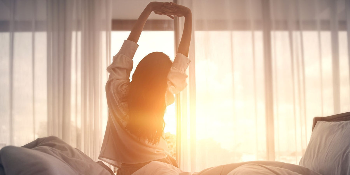 Person stretches arms overhead in a sunlit bedroom by a window.