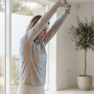 Woman in a light gray long sleeve top stretches arms overhead in a bright, airy home space.
