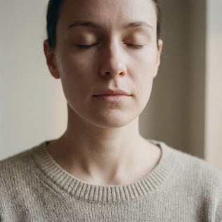 Woman with closed eyes meditates in a calm indoor setting wearing a beige sweater.