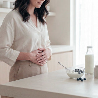Woman in a beige blouse holds her stomach beside yogurt with blueberries and a milk bottle on a light counter.