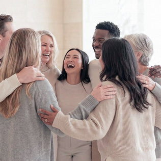 Diverse group of friends in neutral sweaters hug and smile in a bright room.