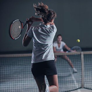 Female tennis player in a white shirt and dark shorts swings a racket on an indoor court as a ball travels toward her with a spectator in the background.