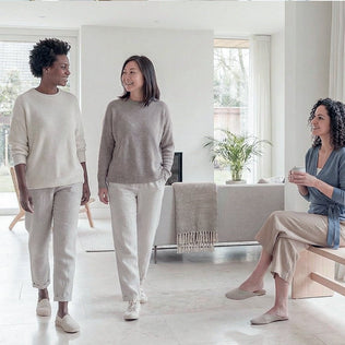 Two women walk and chat in a bright living room while another woman sits on a bench, reaching toward her toes in a seated stretch.