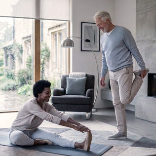 Older couple performing stretches in a modern living room; woman on a mat reaches for her toes as the man stands holding his foot behind him.