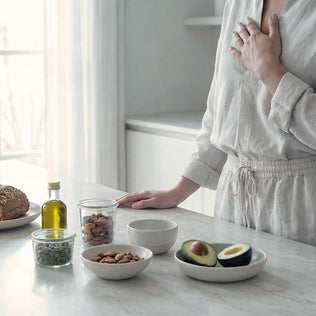Person in light clothing stands at a marble kitchen counter with avocados, nuts and olive oil.
