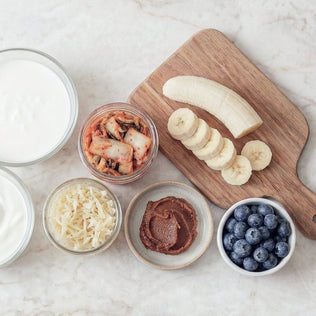 Top down view of probiotic foods on a marble surface including yogurt kimchi coconut flakes nut butter blueberries and banana slices