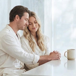 Bright scene of a couple in light clothing leaning on a white counter with hands clasped by a window and a mug on the counter