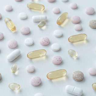 Assorted pills and softgel capsules scattered on a pale surface including white tablets pink speckled pills and amber capsules