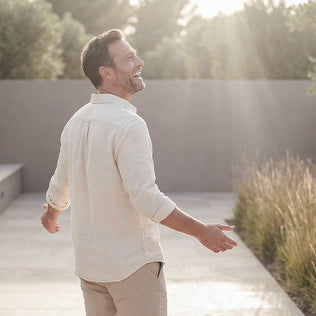 Smiling man in a light linen shirt and beige pants walks outdoors with sunlight streaming and tall grasses in view.