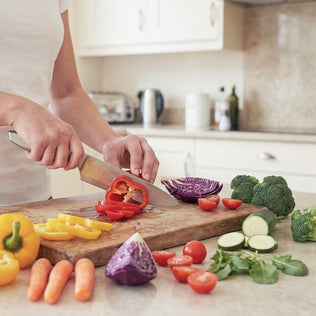 Person chopping peppers on a wooden cutting board with tomatoes cucumbers and broccoli nearby
