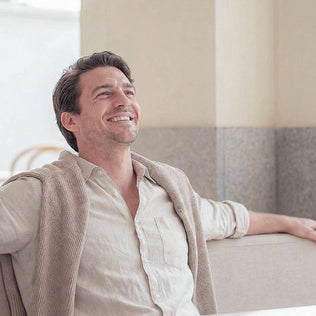 Relaxed man in a beige shirt with a knit cardigan draped over his shoulders smiles in a bright indoor setting